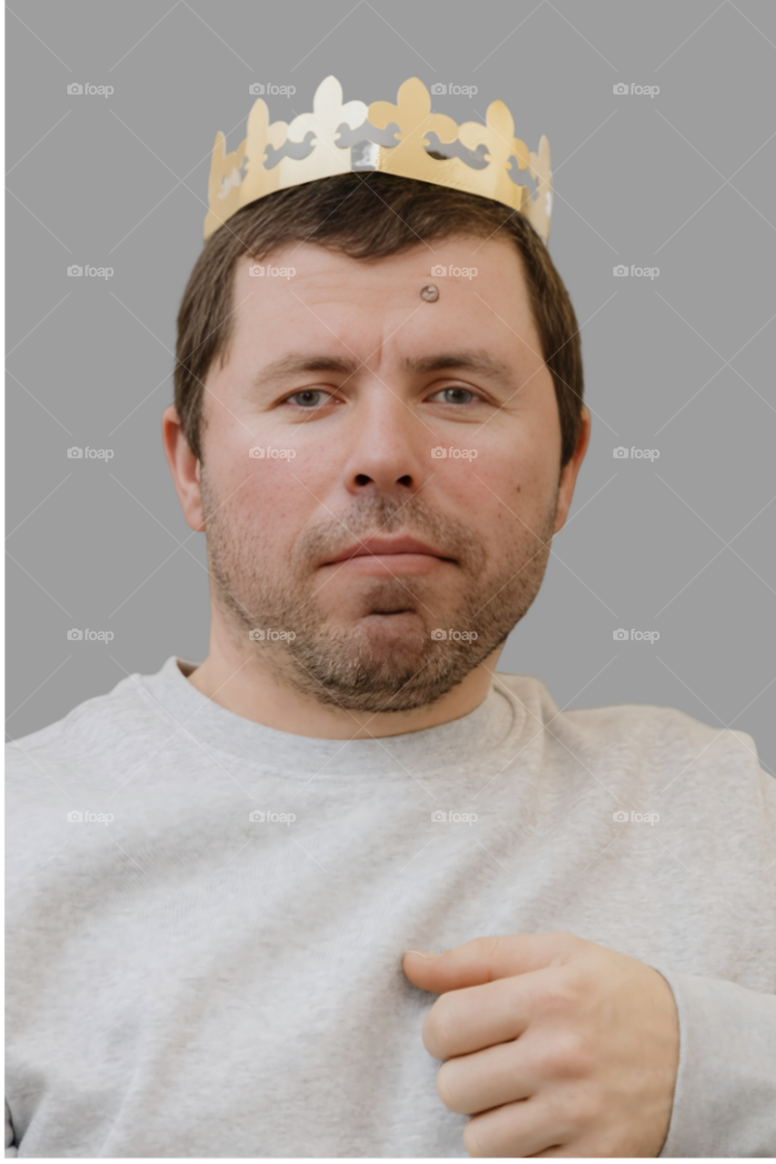 Portrait of a young brunet Caucasian man with a short haircut, stubble and a golden paper crown on his head looking at the camera with a smile against a white wall in a room in the daytime, close-up view from the side.