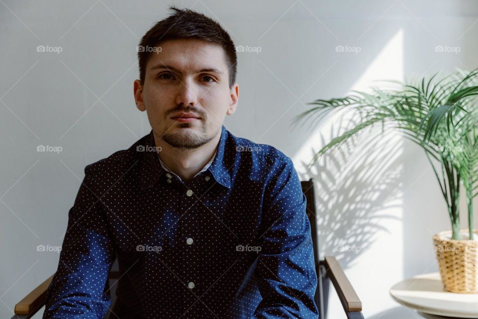 Portrait of one beautiful Caucasian young guy in a blue shirt with a smile looks at the camera and sits in a chair in a room on a summer day, side view close-up.