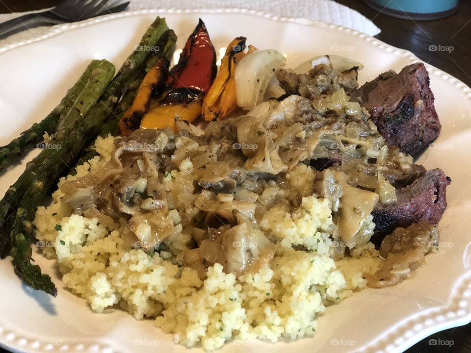 Grilled venison and morel wild mushrooms over a bed of Parmesan couscous, paired with grilled asparagus, peppers and onions. All served on a white dish.