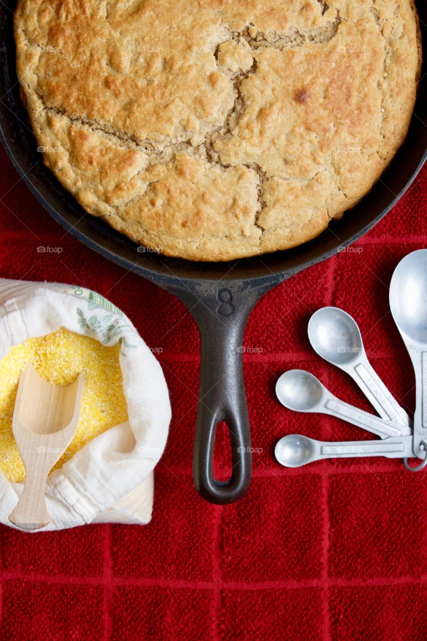 Flat lay of sourdough cornbread in a cast iron skillet, cornmeal and wooden scoop in a flour sack, vintage measuring spoons on a red kitchen towel