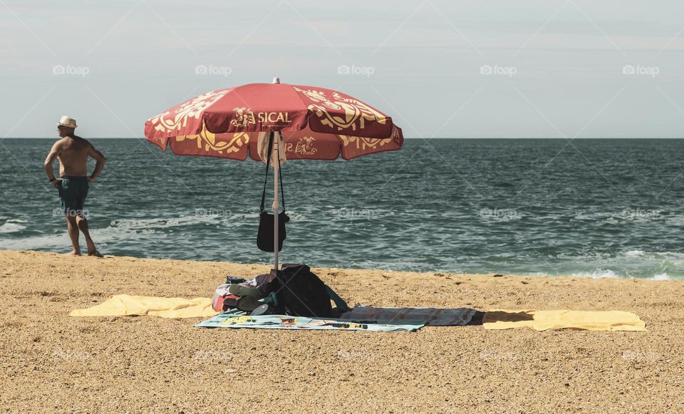 A parasol shields a pile on personal belongs from the sun, as a man considers a swim at Praia da Nazaré, Portugal 