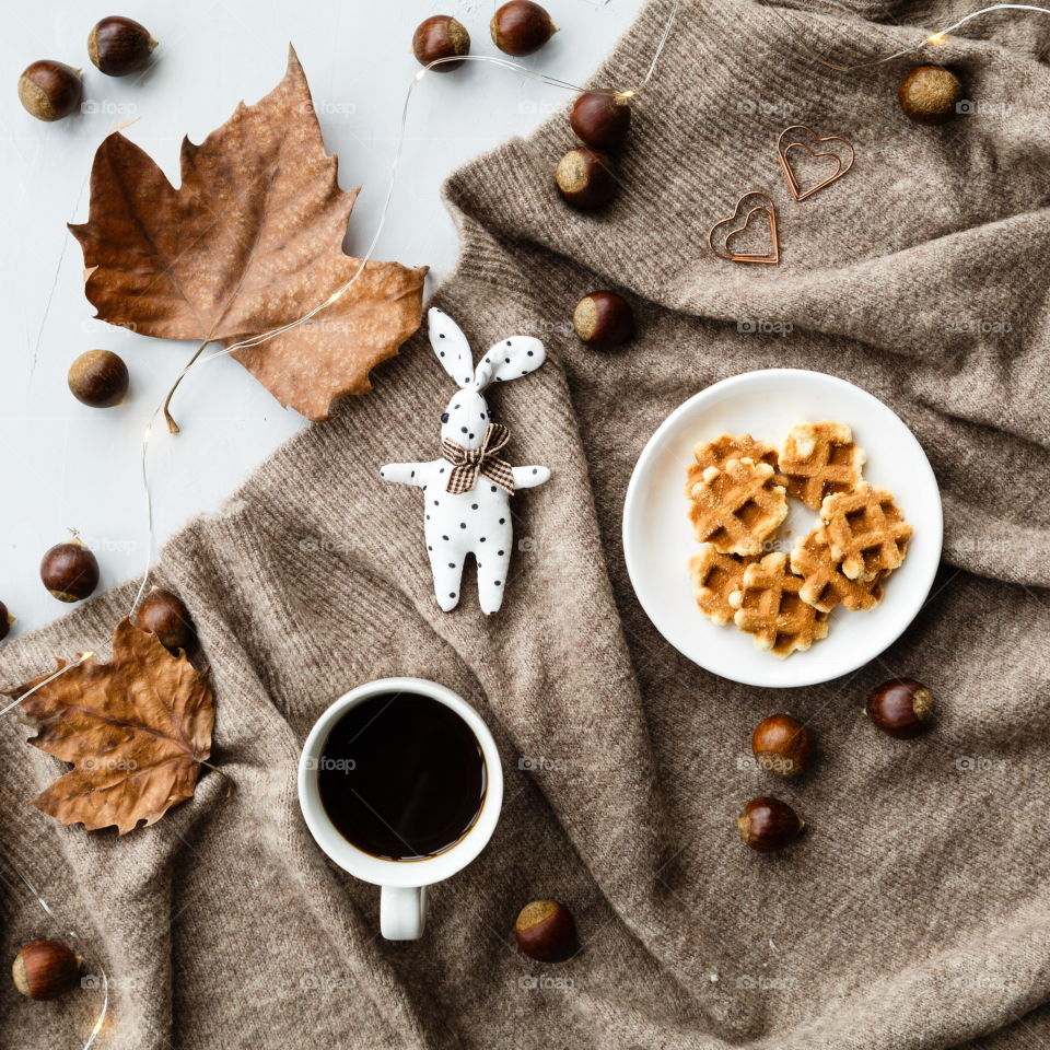 Flatlay items: cup of coffee, plate with cookies, chestnuts, maple leaves, rabbit toy, garland, knitted sweater brown color