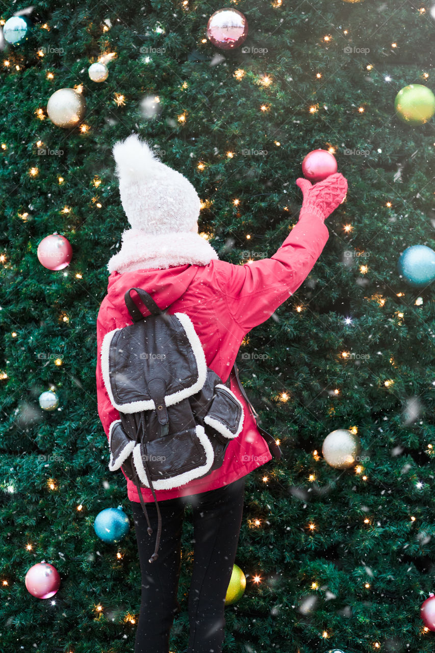 Young girl standing at front of big Christmas tree decorated with colorful balls. Teenager girl is wearing warm clothes, scarf and wool cap on a cold winter day