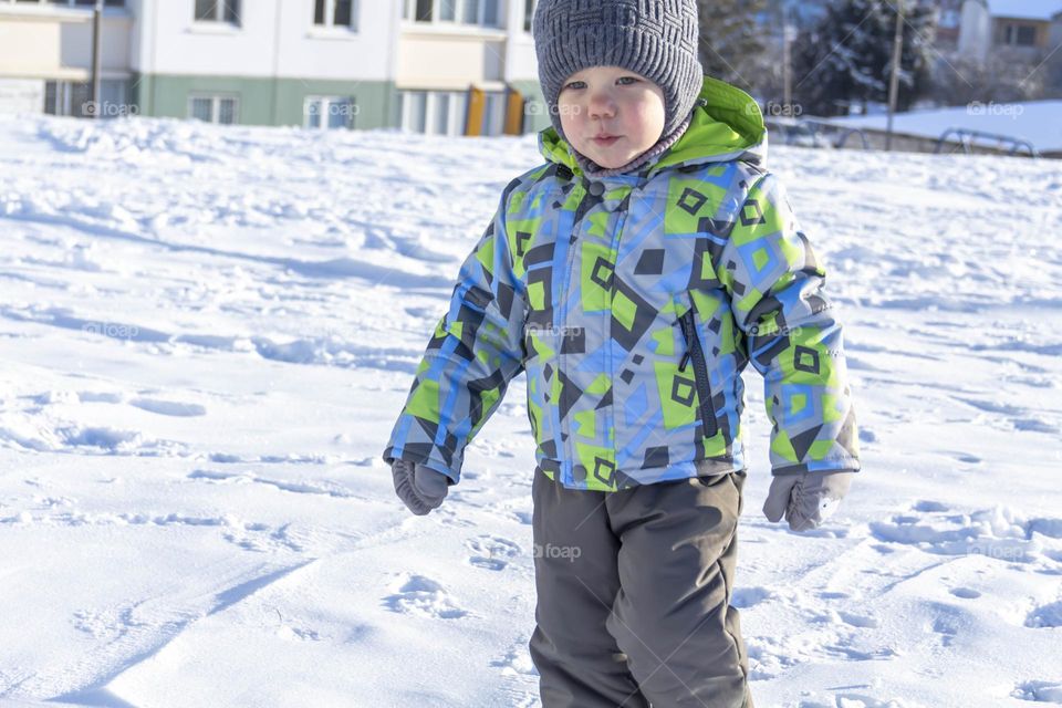A child with a serious expression on his face in winter clothes jackets, pants, hat and boots in winter on the white snow on the street and in the park in nature plays winter fun.