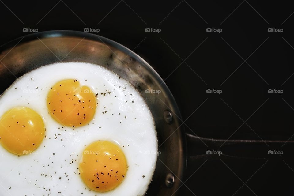 Three Sunnyside up eggs with pepper cooking in a frying pan with a black background
