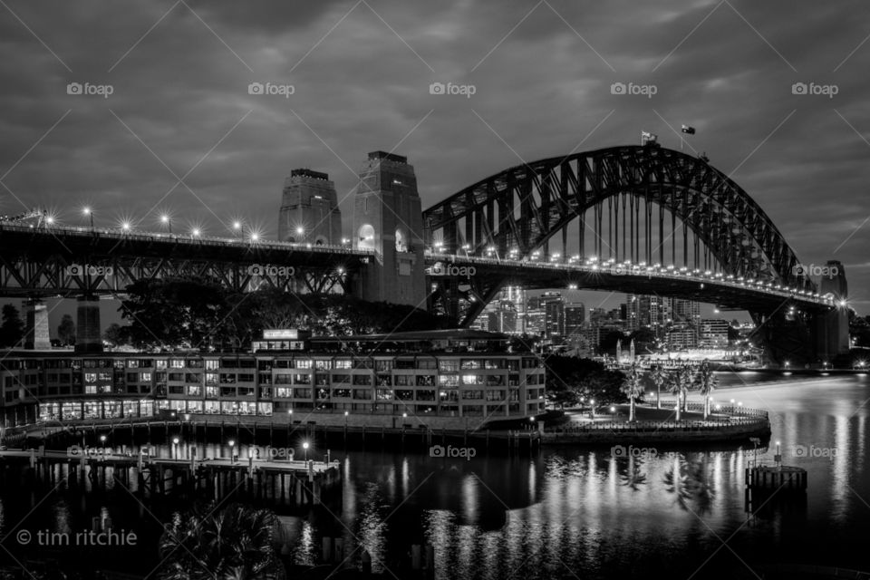 Under pre dawn clouds, Sydney’s Harbour Bridge lights reflect off the water while Campbell’s Cove lays asleep