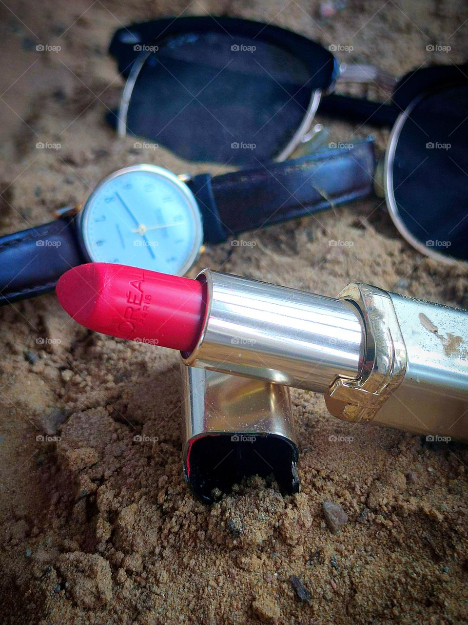 There is a lipstick cap on the sand with a tube of open red lipstick leaning against it. In the background, a wristwatch and sunglasses lie on the sand.