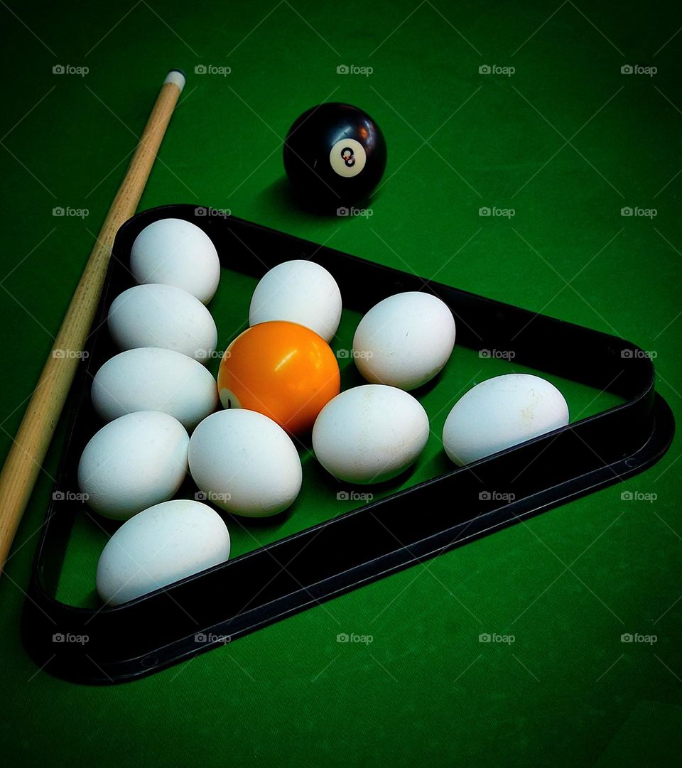 There is a black triangle for pyramids on the green billiard table.In the billiard triangle lie white chicken eggs,among which lies a yellow billiard ball as a contrast.Also on the billiard table is a wooden cue and a black billiard ball.White colour