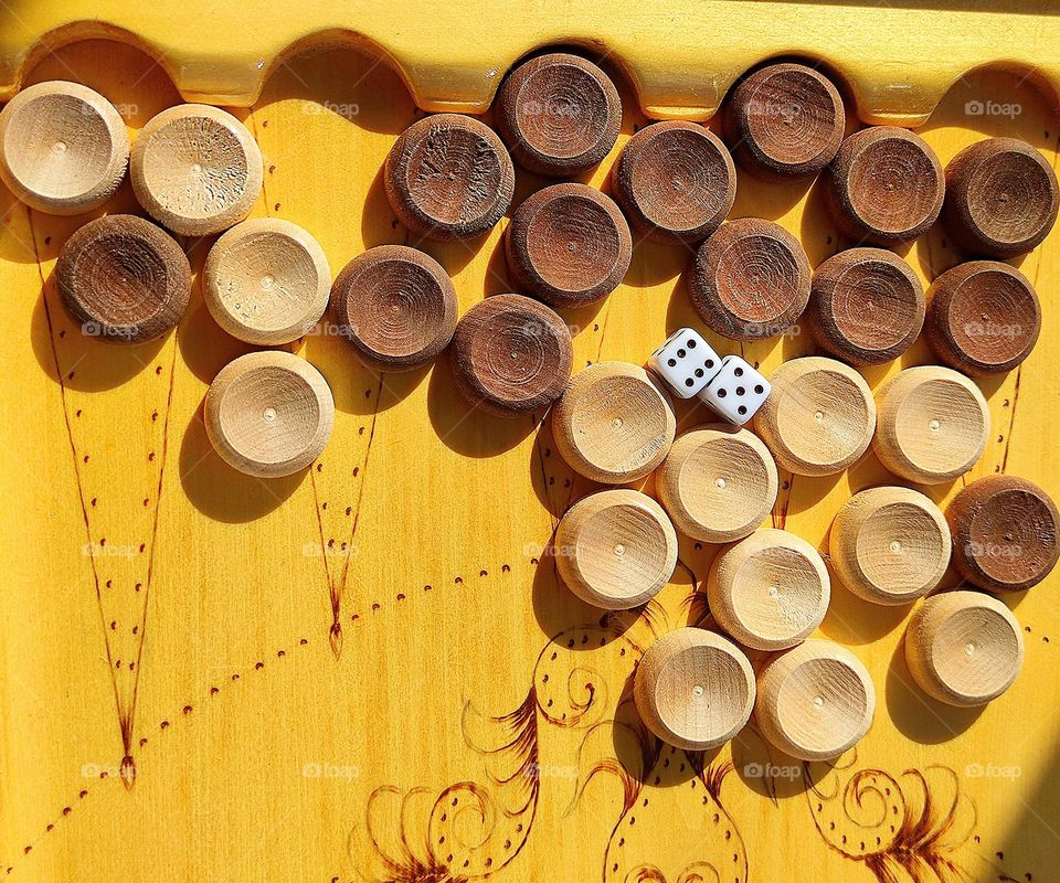 Circles. Backgammon game. On the wooden surface of the board are wooden round white and black chips. Among the chips are two white dice (cubes) with black numbers in the form of holes.