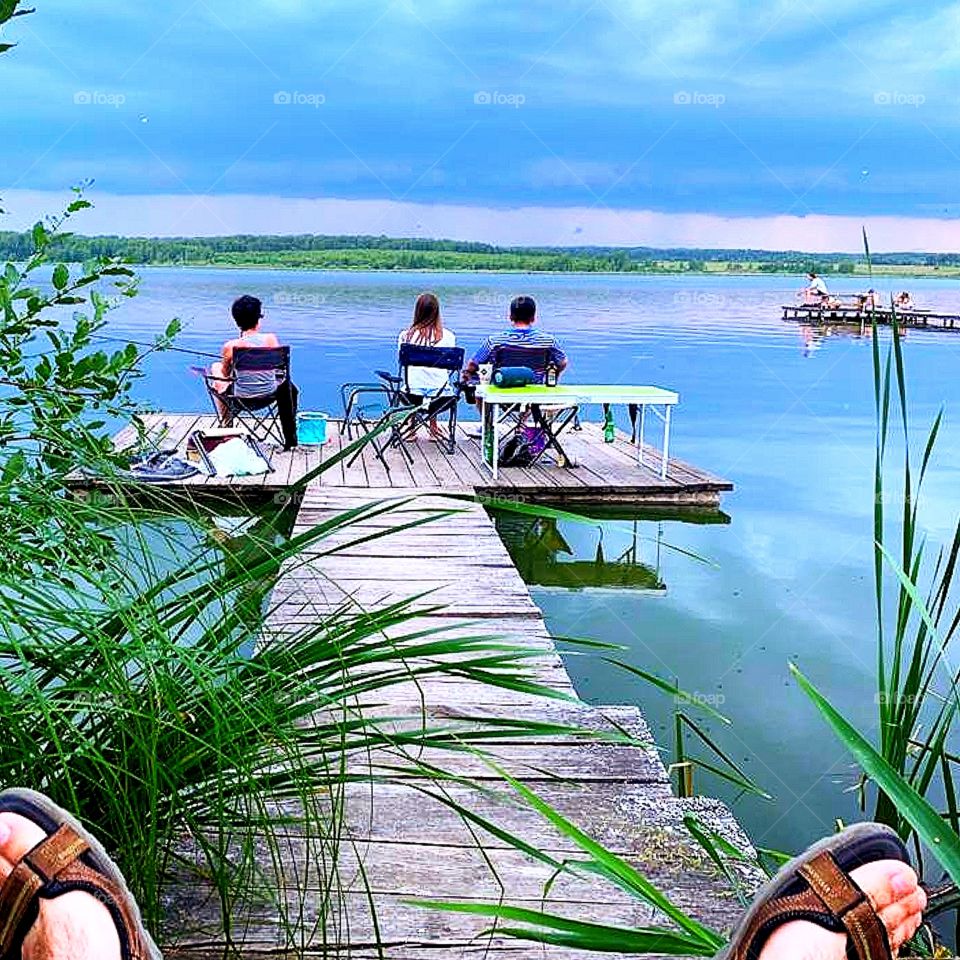 Three fishermen sit on a wooden bridge and fish.  In the foreground are bare feet in summer slippers.