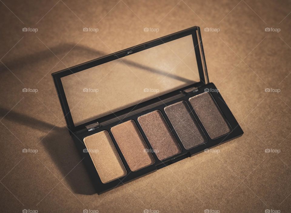 One eyeshadow palette in nude shades with an open transparent lid lies diagonally on a dark brown background, close-up top view. Female concept cosmetics,beauty.