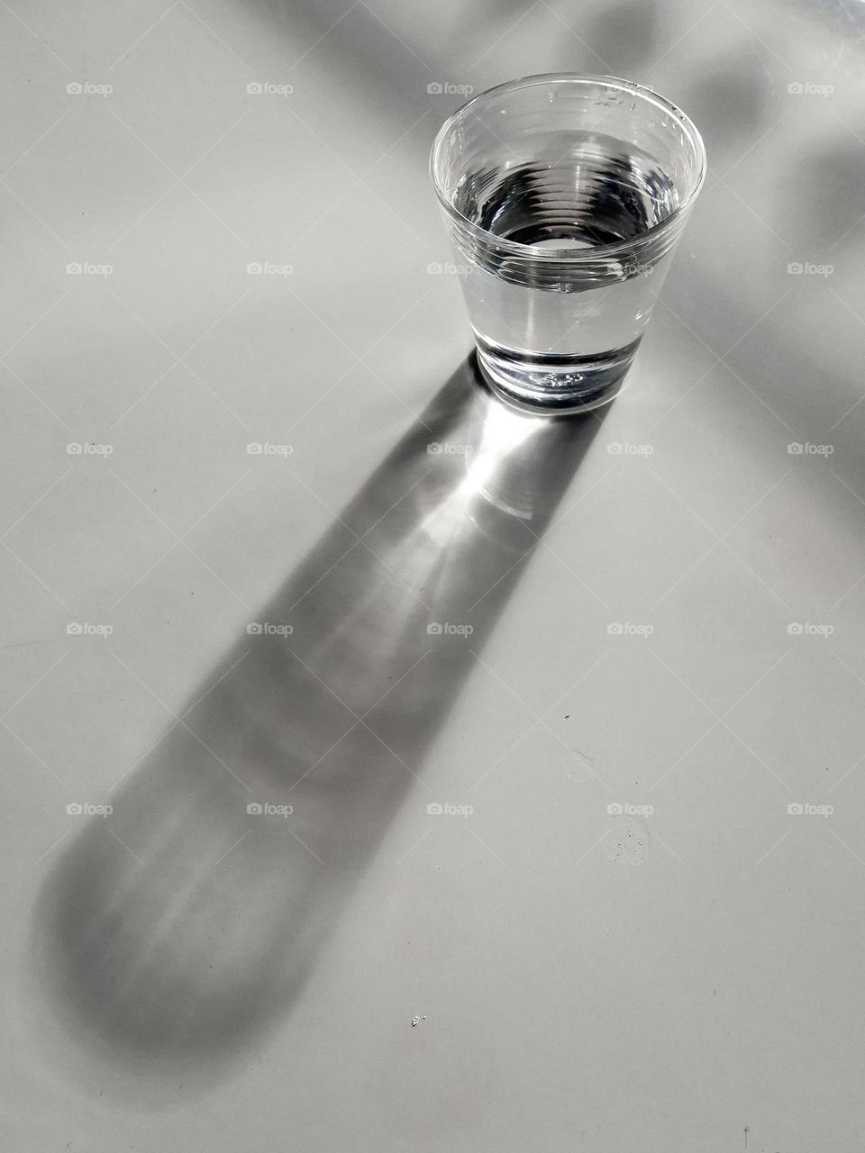A glass with clean clear water and sharp shadows stands on a white wood table. top view of glass with water on white table.