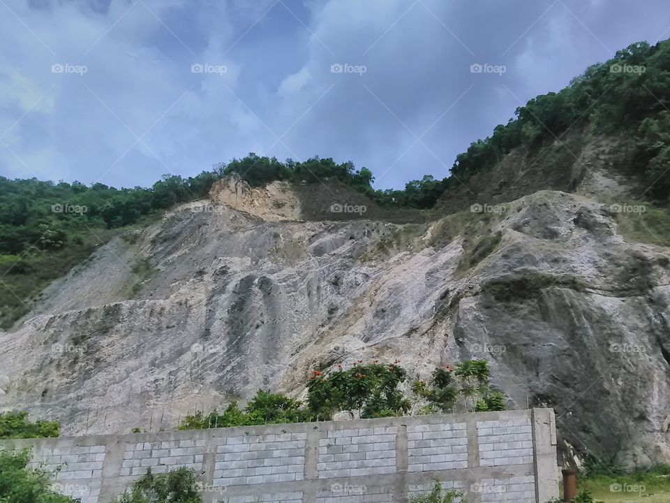 Great wall. Huge limestones forming a mountainous terrain. Concrete retaining wall and green shrubs, with blue and white clouds.
