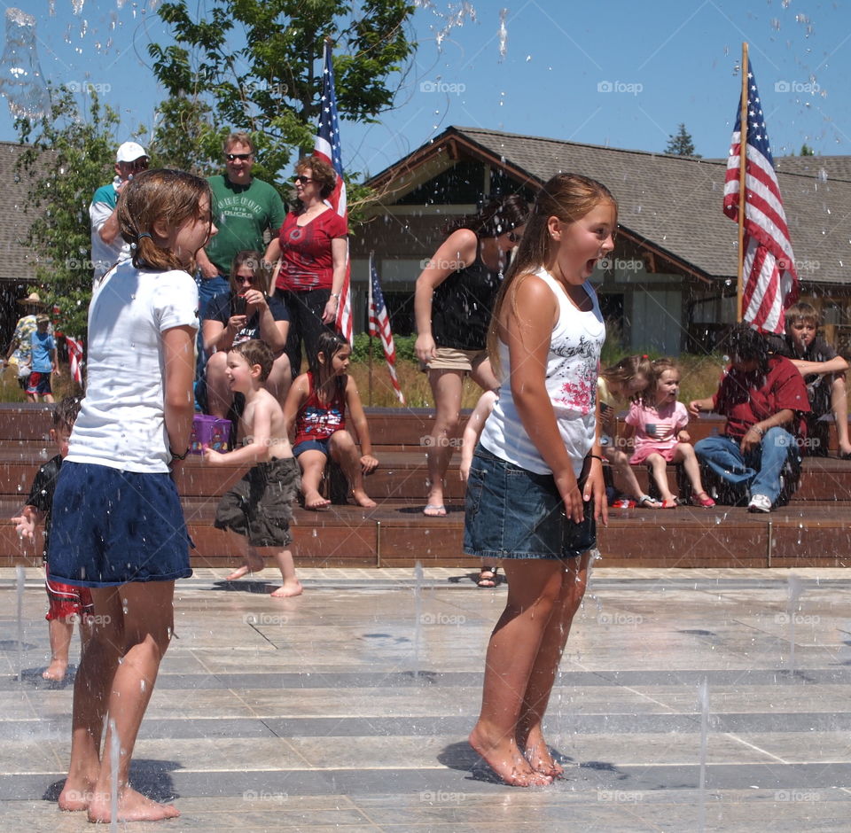 Two young girls having fun from the surprise of water jets at the water play feature for kids at Centennial Park in Redmond in Central Oregon on a sunny summer afternoon. 