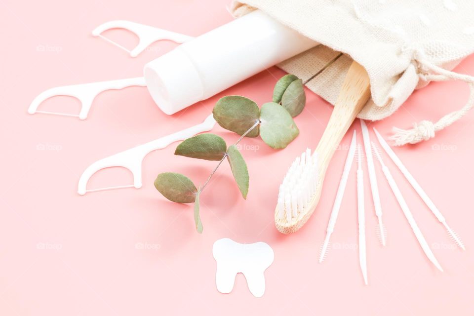 One white tube of toothpaste with a bamboo toothbrush, a cut out paper tooth, tooth sticks and a branch of greenery are scattered from a beige burlap bag with an openwork insert with drawstrings on a pink background, close-up side view. Dental health