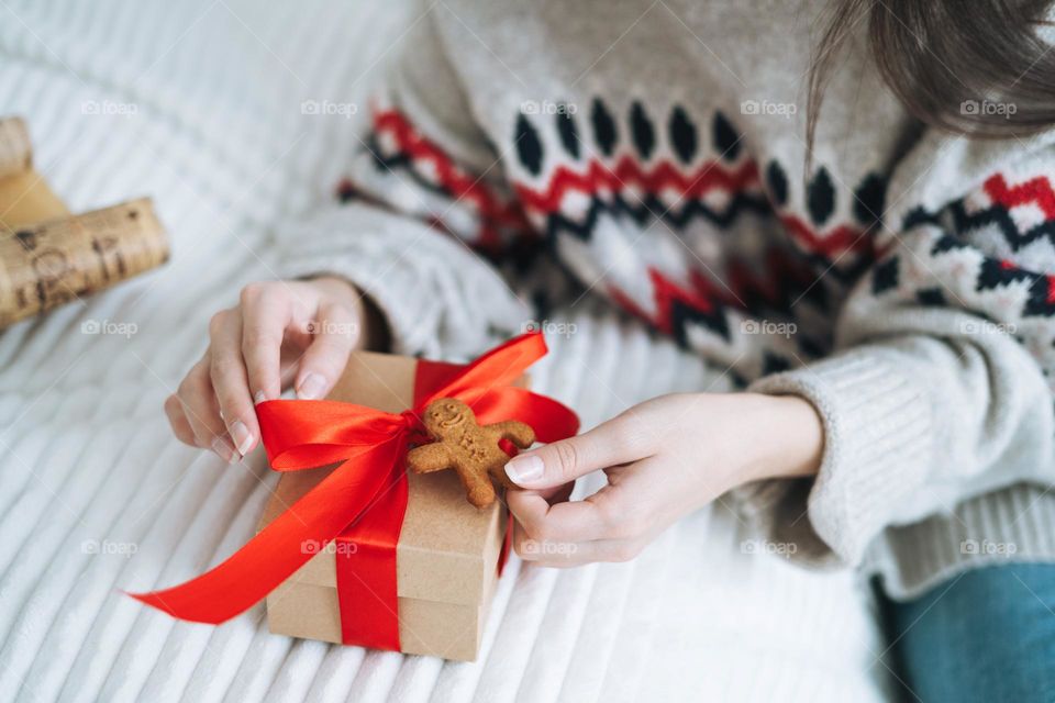 Young woman in cozy sweater with gift box with red ribbon in room with Christmas tree at home