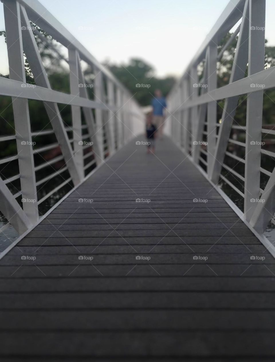 White metal bridge with wooden walkway with two pedestrians in the background. Green trees and a clear blue sky accent the picture. 