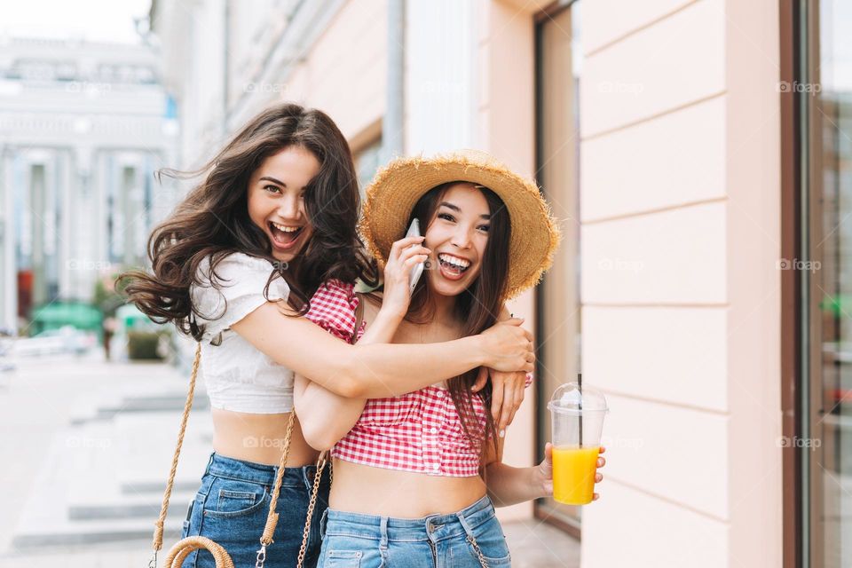 Happy smiling beautiful brunette young women friends in summer clothes with juice in hands having fun on summer city street