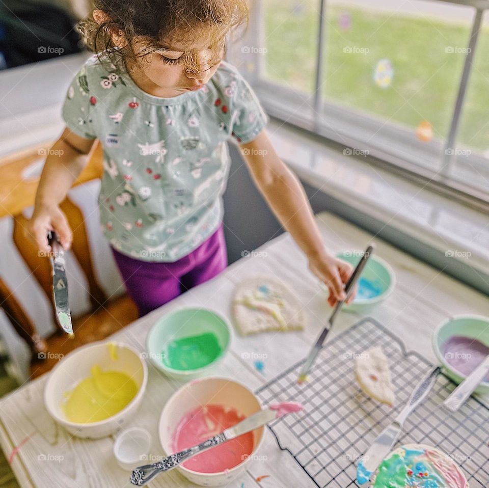 Toddler girl frosts Easter sugar cookies, making homemade sugar cookies with icing for Easter, Easter traditions with children, making cookies with kids, baking homemade cookies with kids, colorful cookies made by a toddler 