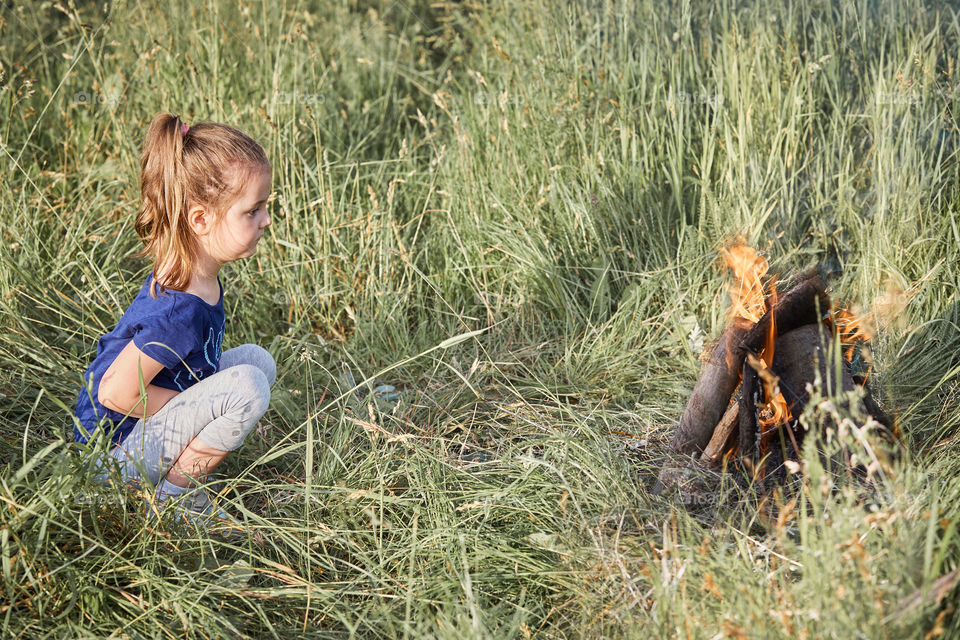 Little girl looking at campfire sitting in a grass. Candid people, real moments, authentic situations