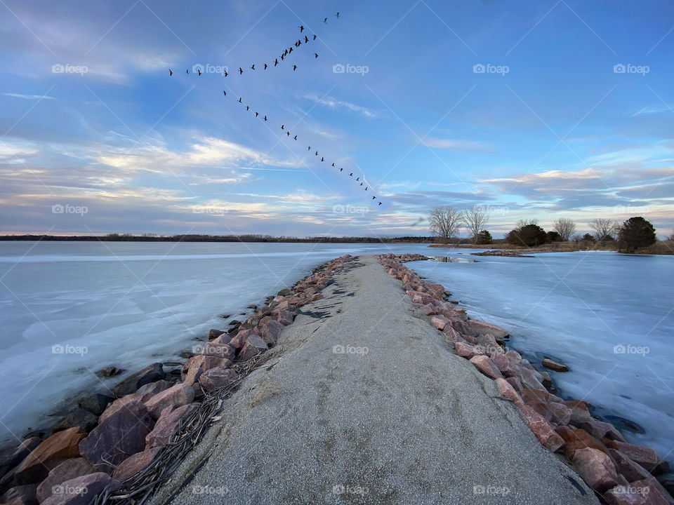 A rocky jetty on a frozen lake with geese flying in a V formation above the lake