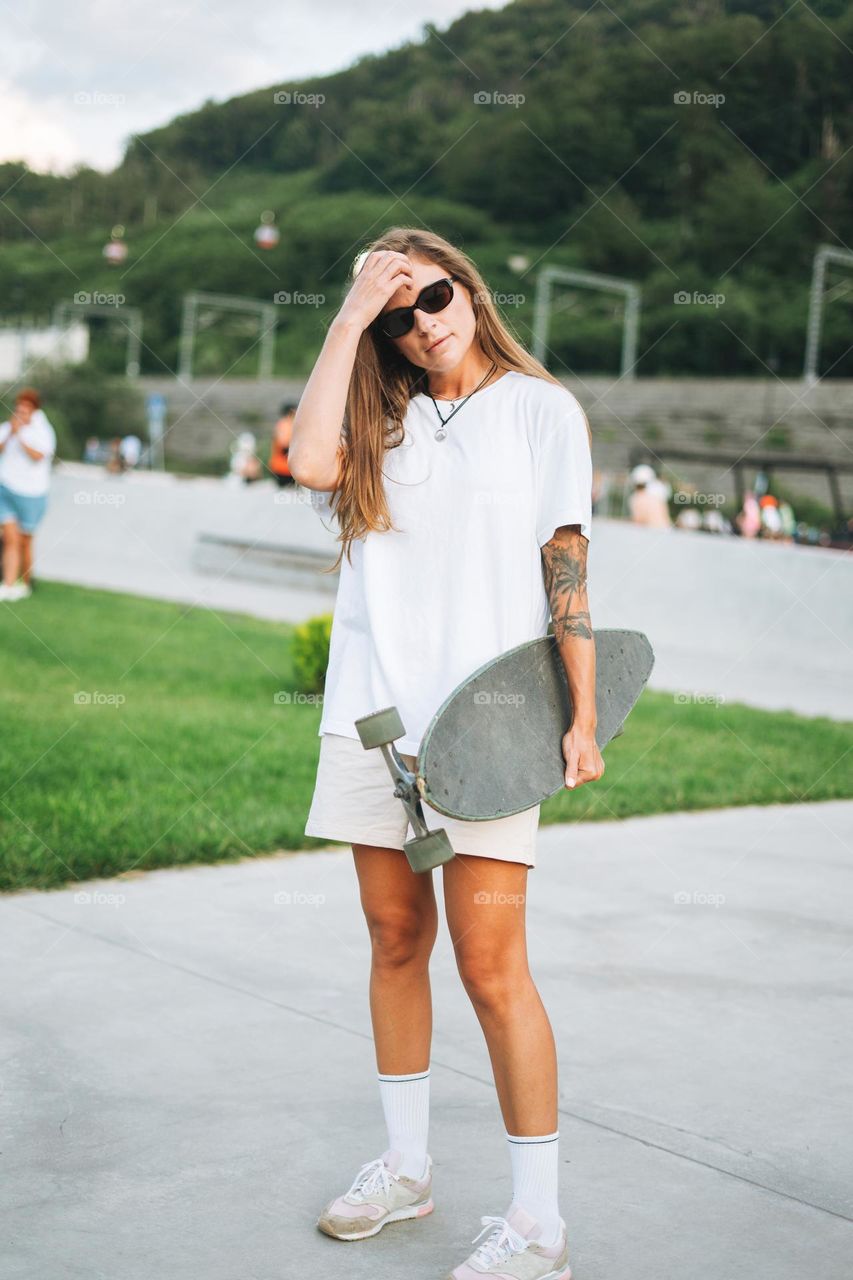Slim young woman with long blonde hair in light sports clothes with longboard in the outdoor skatepark at sunset