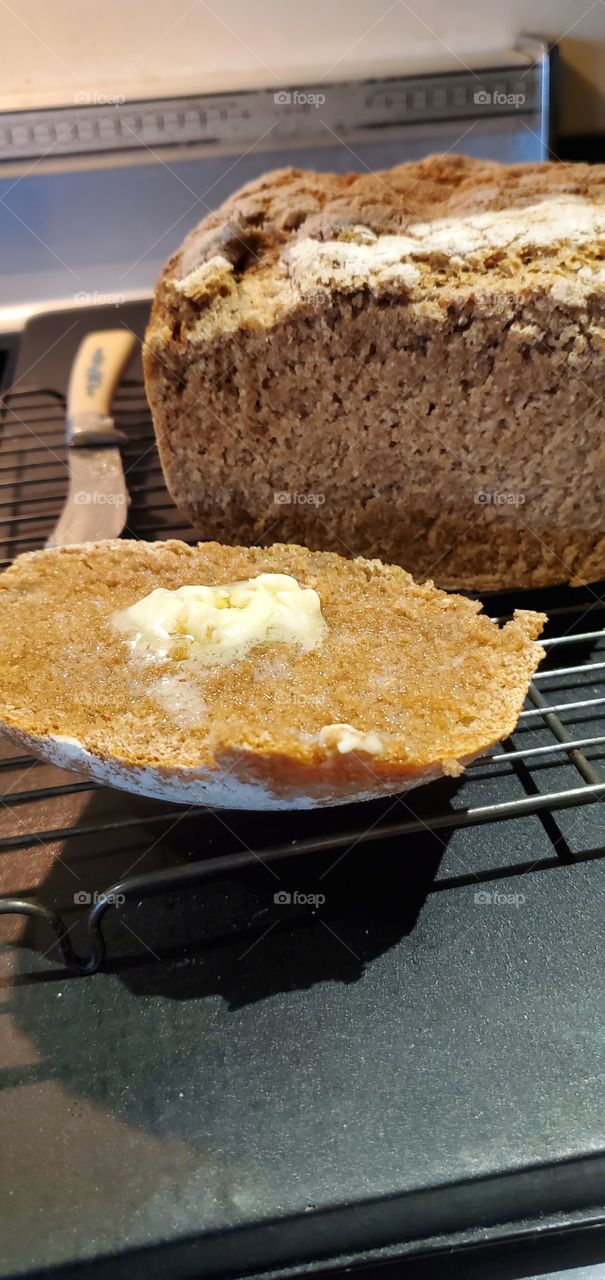 Hot buttered slice of homemade stone ground whole wheat bread. Bread sitting on cooling rack with butter knife next to it.