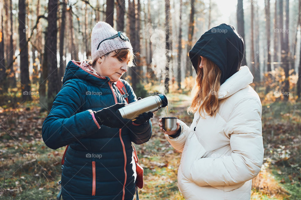 Mother and daughter with backpack having break during autumn trip pouring a hot drink from thermos flask on autumn cold day. Active women wandering in a forest actively spending time