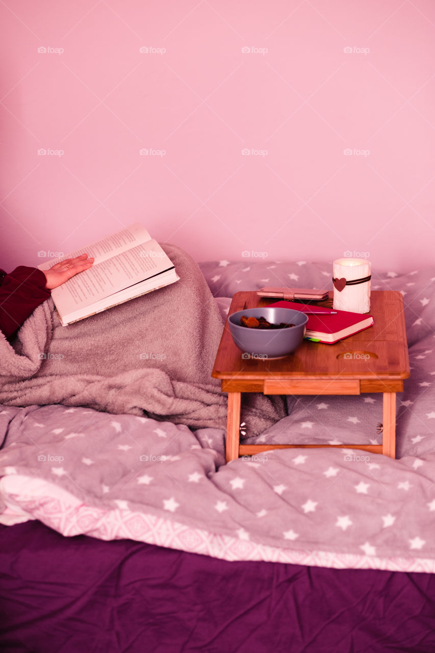 Woman enjoying the reading a book and drinking coffee at home. Young woman sitting in bed, wrapped in blanket, holding book, relaxing at home. Candid people, real moments, authentic situations