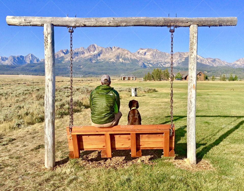 A man and his dog sitting on a park swing, taking in the views of the beautiful Sawtooth Mountains in Stanley, Idaho.