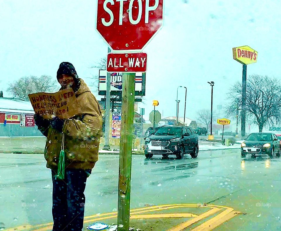 Homeless man on street holding sign next to Stop Sign in traffic 