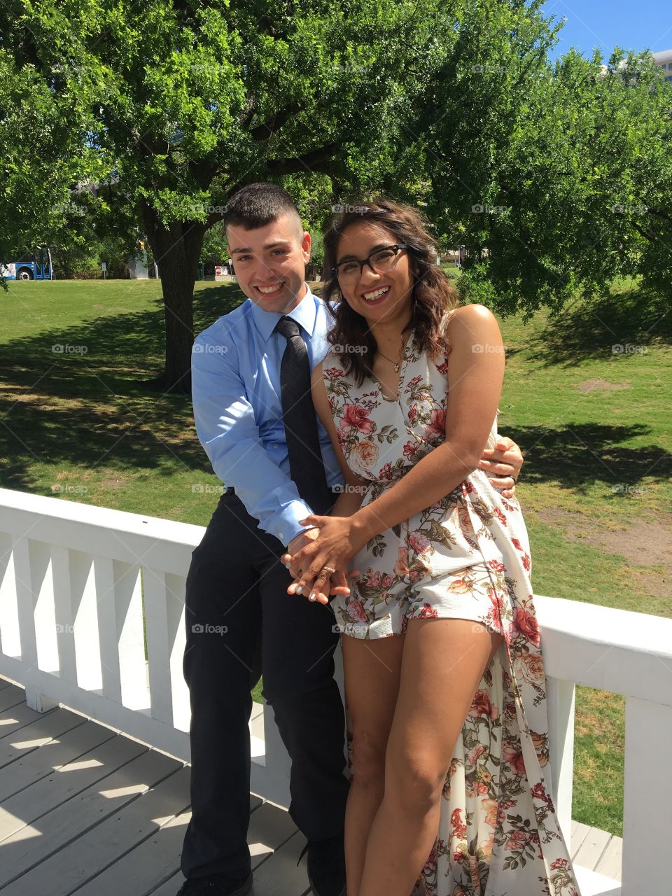 A newlywed couple sits on the ledge of a white gazebo. She wears a floral romper/dress, he wears a powder blue button-up. He has his one arm wrapped around her, the other is holding her hand. Both look at the camera with loving smiles. 