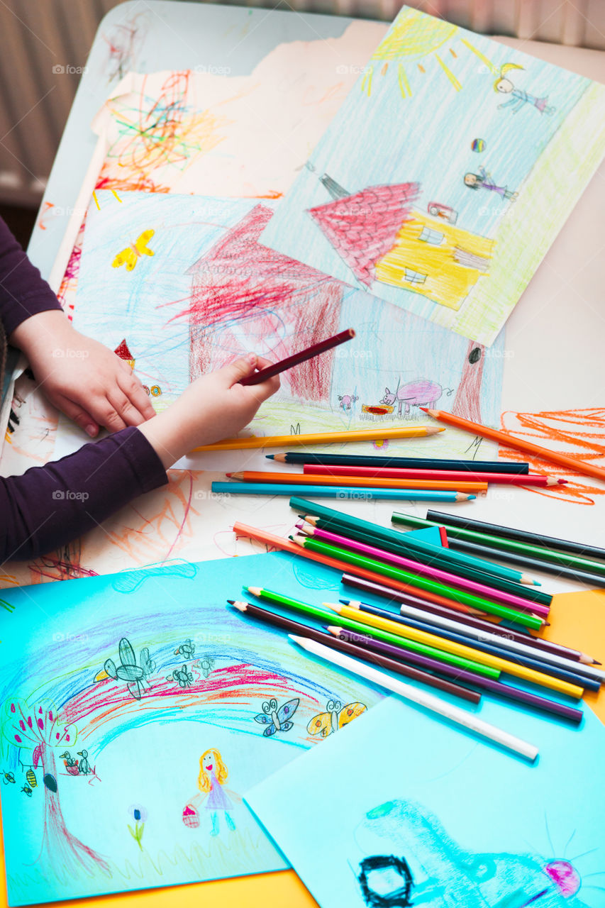 Pencil crayons scattered on desktop filled with colorful childlike drawings of playing children, house, car. Child hand holding pencil. Photo from above