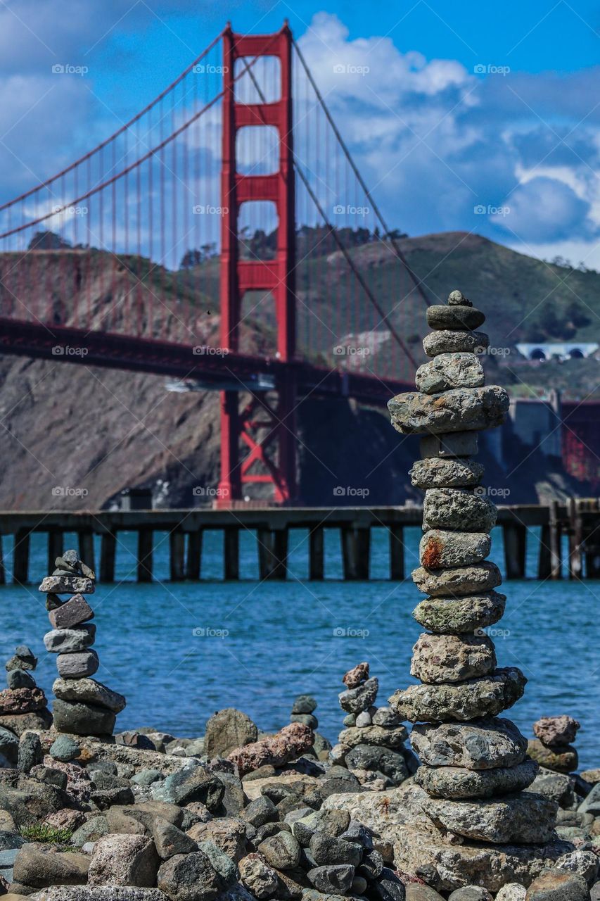 Golden Gate Bridge with rock stacks forming little towers in the foreground, with a beautiful cloudy blue sky and the San Francisco Bay chilling.