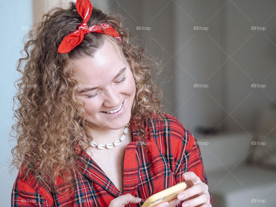 Young beautiful woman with curly hair in plaid shirt using mobile phone and smiling 