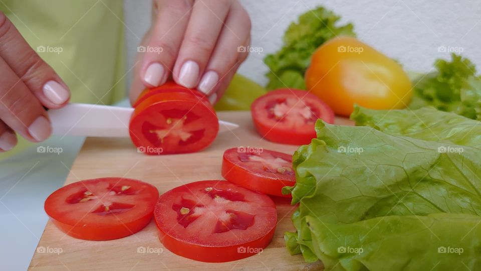 Cooking salad at home. Cutting vegetables. Wooden cutting board, ceramic knife, tomatoes, yellow pepper, lettuce.