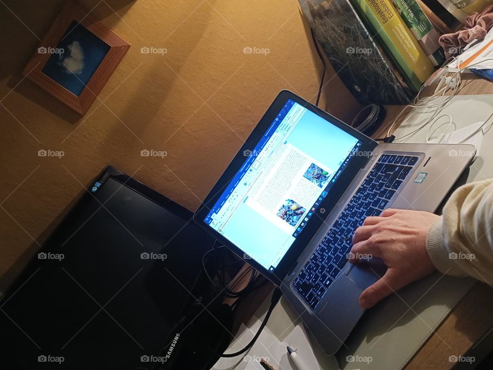 Close-up of a hand writing on a laptop on a desk filled with a black screen and books, at the background a picture of a cloud heart on a yellow painted wall