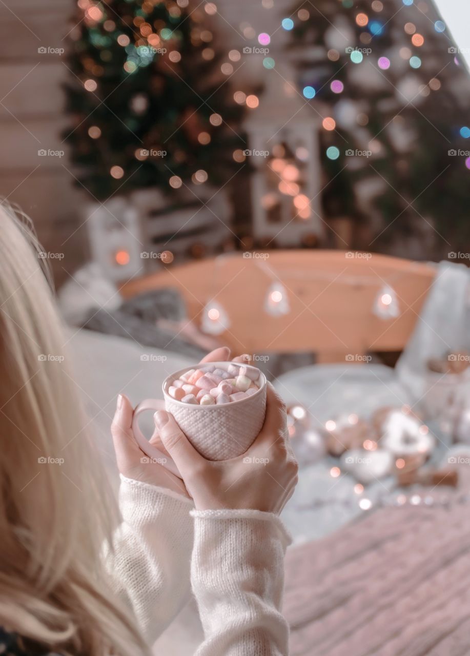 Cup of cocoa with marshmallows in the hands of a girl in a room decorated for Christmas