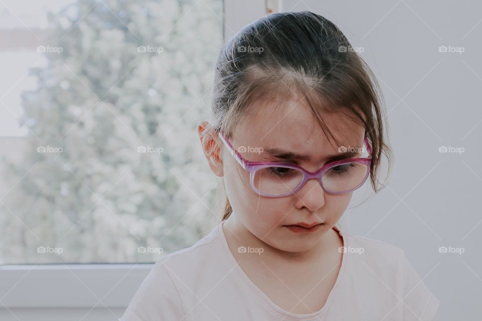 Portrait of a beautiful caucasian brunette girl in glasses with a sad and offended emotion to tears on her face, stands in a room opposite the window, close-up side view. Sad people concept.