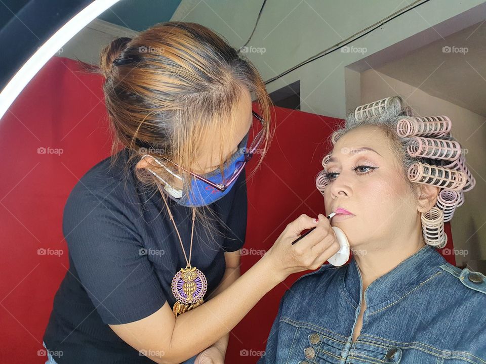 A make up artist wearing a mask is applying lipstick on a model's lips in a photo studio