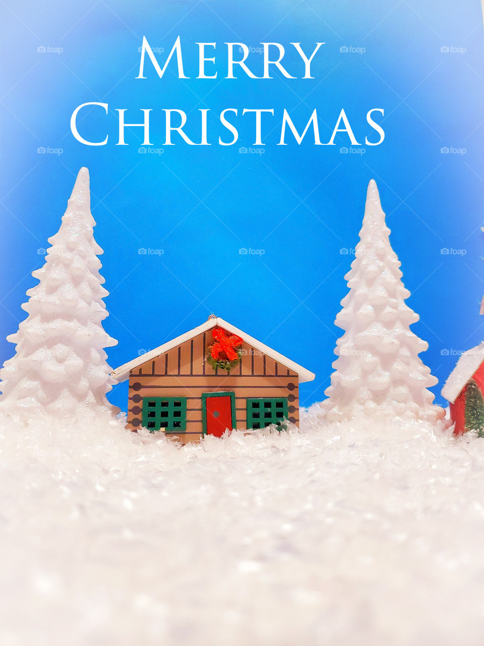 Winter miniature scene of a log cabin and two snow covered pine trees surrounded by snow with the greeting Merry Christmas in white.