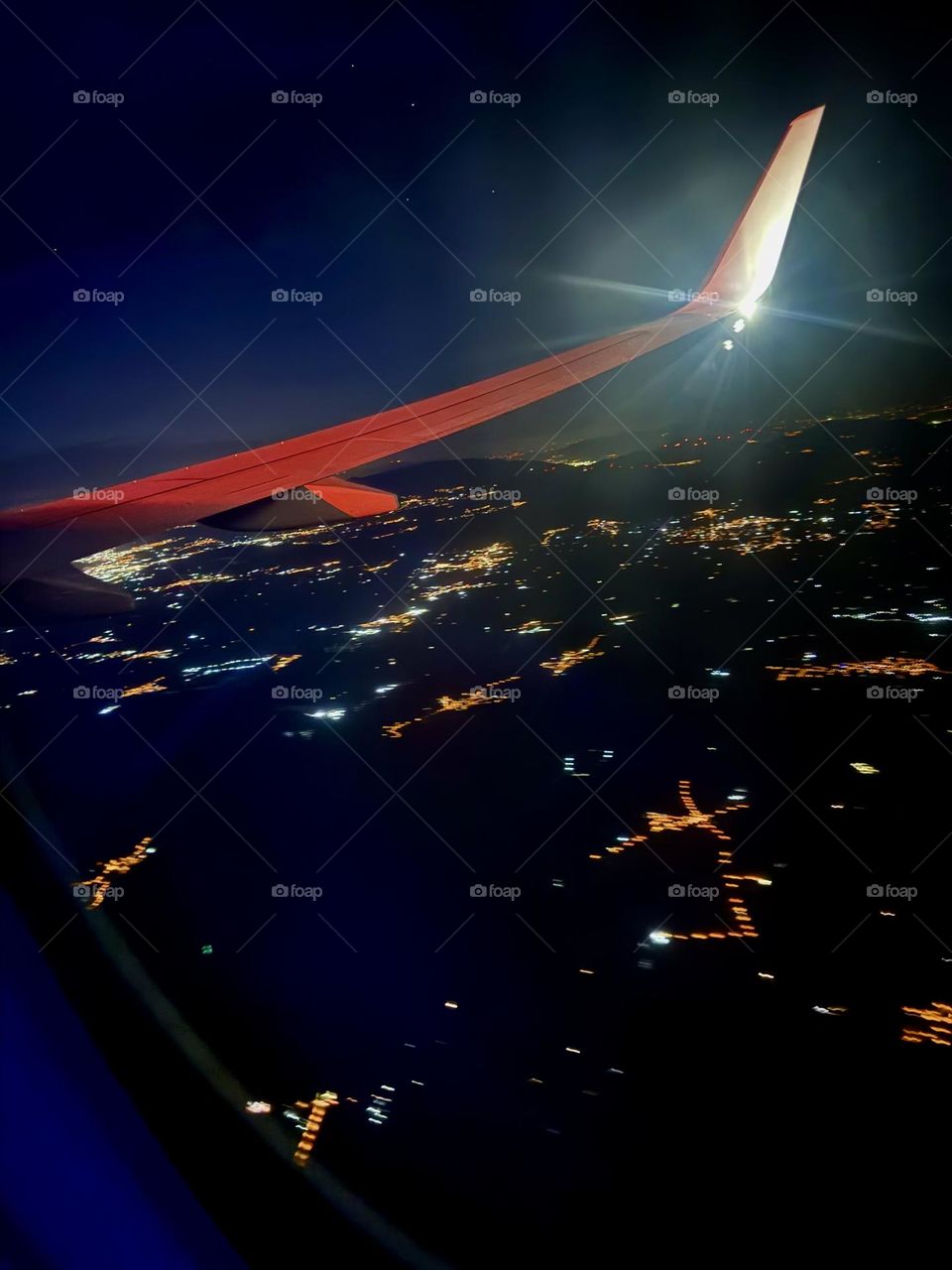 На фото виден фрагмент крыла самолета, который освещен тусклым светом иллюминаторов. Внизу, под крылом, раскинулся ночной город, мерцающий тысячами огней