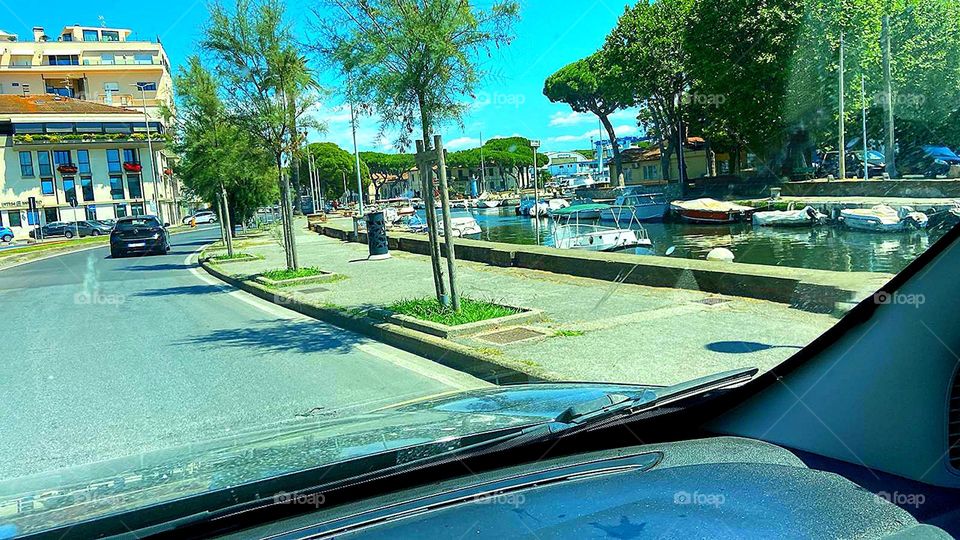 View from the windshield.  A road along which there are green trees, a water channel in which there are boats and small yachts