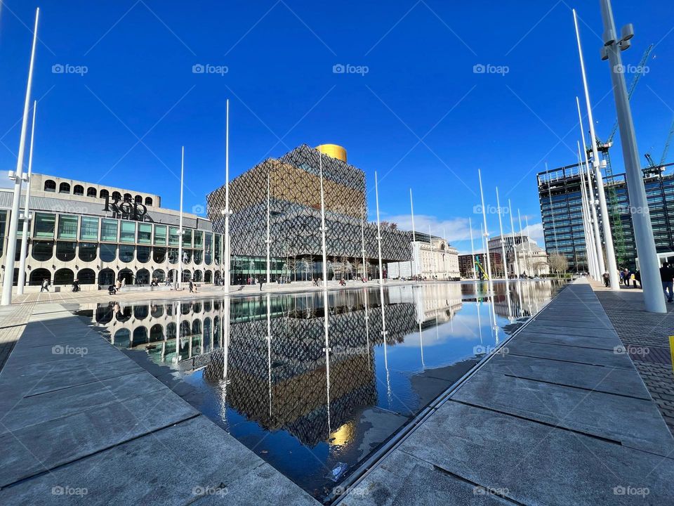 Reflection of Birmingham’s library in the water by day with blue sky and sunshine.