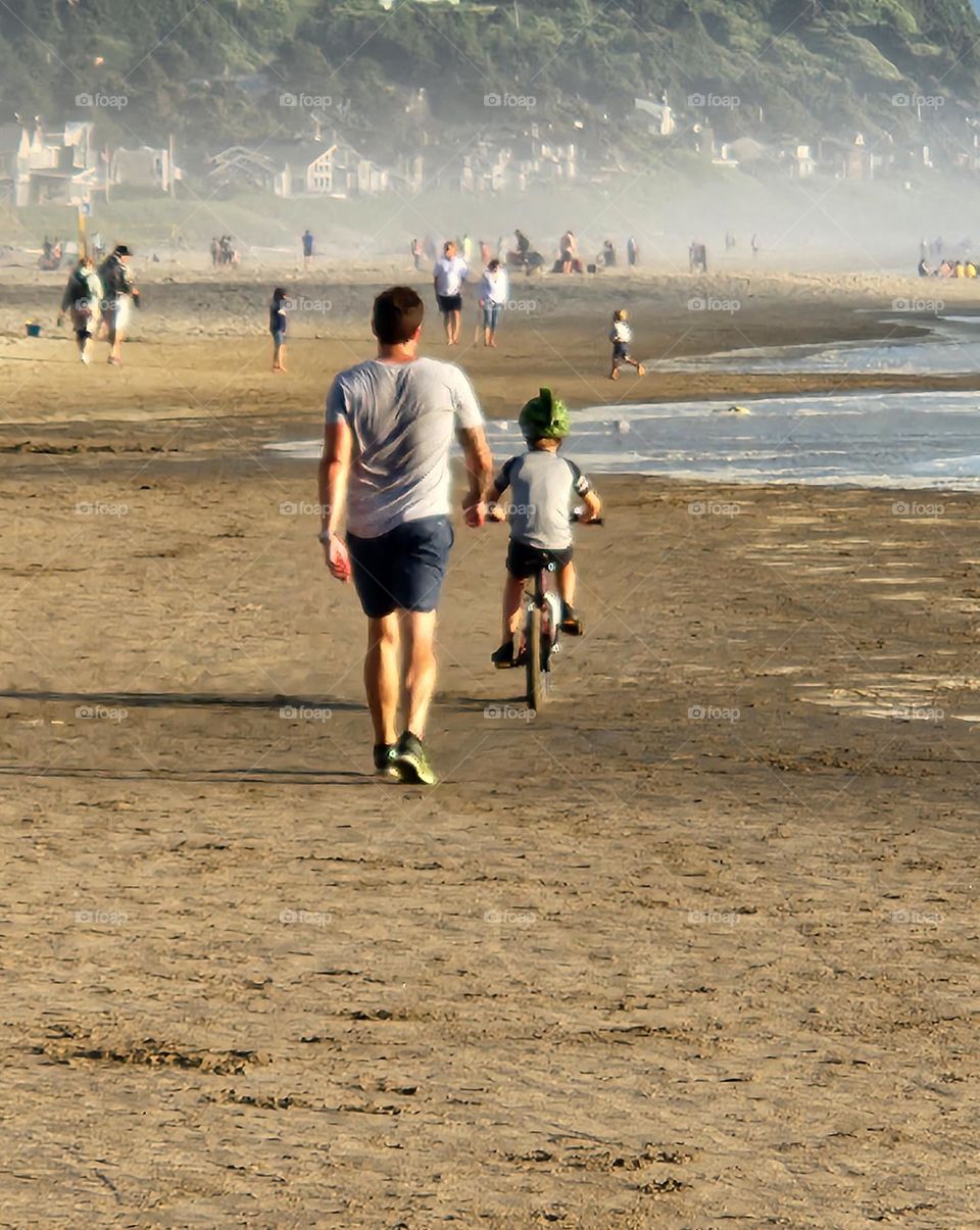 man walking alongside his son bicycling across the sand on a sunny Summer evening in Oregon