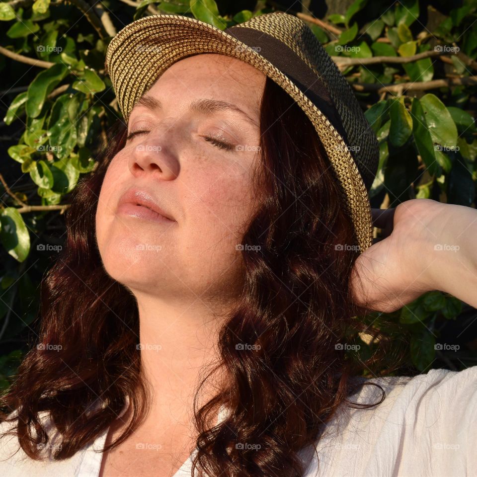 Women with natural and minimal makeup standing outdoors outside with wet hair. Women with red hair and green eyes standing next to tree taking a selfie. Sunlight shine on women hair and skin and making shadows. Women wearing a hat.