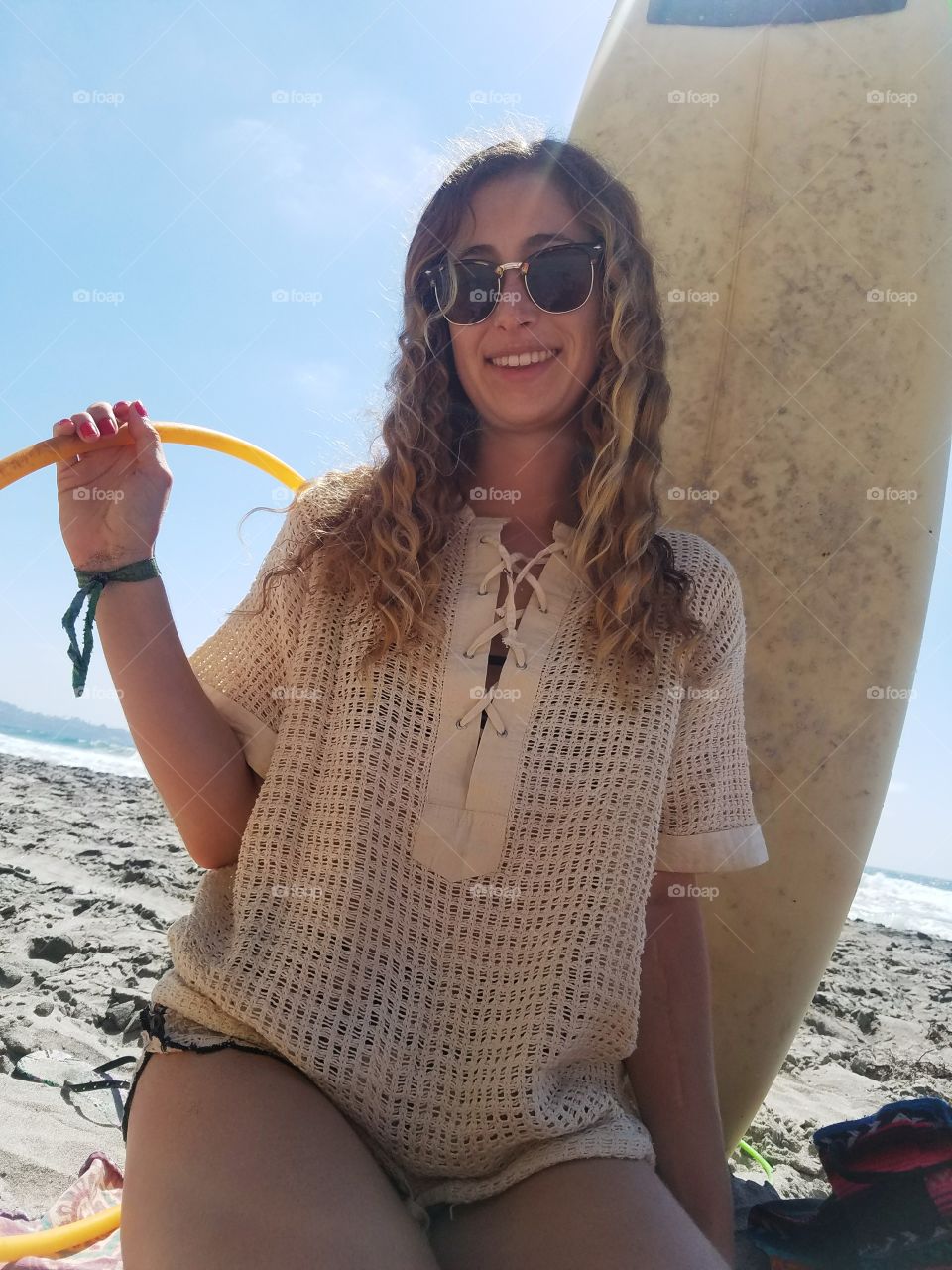 a girl with sunglasses and a hulahoop sitting in front of a surfboard on the beach