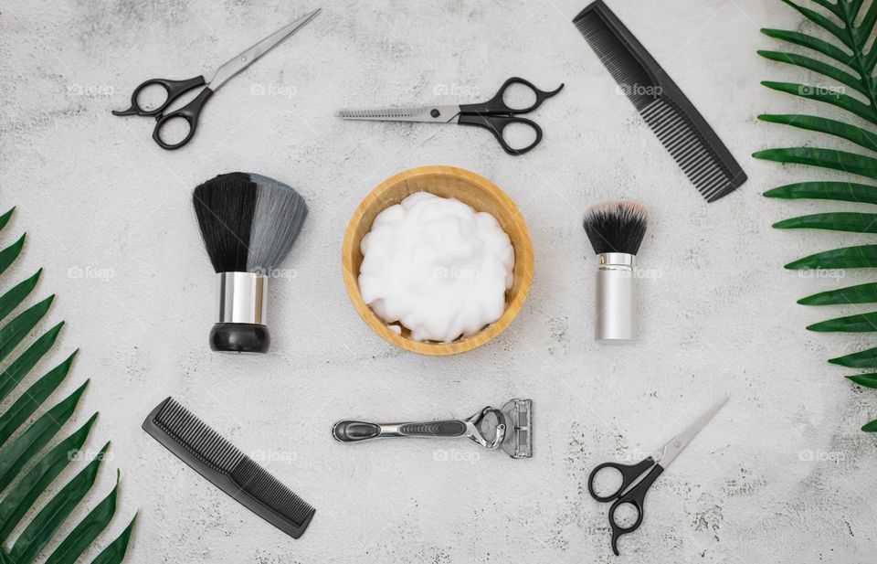 Wooden bowl with shaving foam, and around it are scissors, combs, razor and brushes with a natural pile for shaving on a light gray stone background , flat lay. The concept of barbershop men, shaving men, beard care.