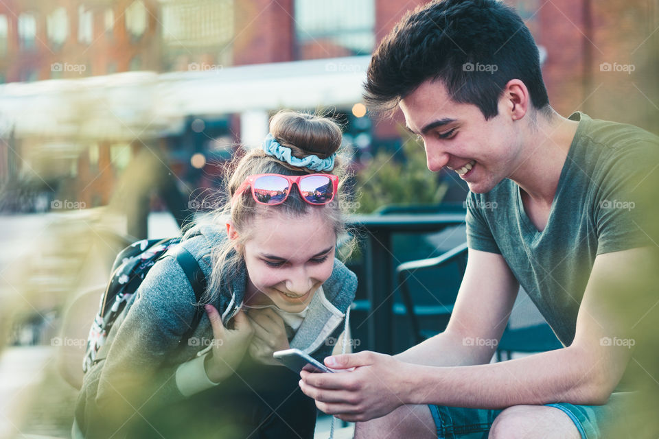 Couple of friends, teenage girl and boy, having fun with smartphones, sitting in center of town, spending time together