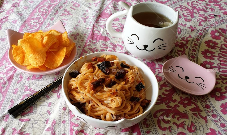 A plate of crisps (chips), a bowl of fried noodles, and a cup of herbal tea on a table. Cat themed tableware.